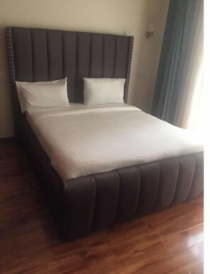 Uma cama com cabeceira escura e duas almofadas, chão de madeira e parede em tom claro.