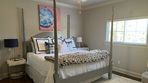 Ein Bett mit Baldachin, ausgestattet mit einer weißen Decke und einer beigefarbenen Tagesdecke. Ein Nachttisch mit Lampe und einer kleinen Dekoration steht daneben. Darüber hängt ein Gemälde an der Wand.