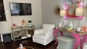 Ein Wohnzimmer mit einem weißen Sessel, einem Holztisch mit rosa Akzenten, einem Glastisch mit bunten Verzierungen und einem flachbildfernseher an der Wand.