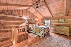 A log cabin bedroom with a bed, nightstands, a chest of drawers, a ceiling fan, and a large window. - 'Turkey Hollow Cabin' w/ Stunning Open Views! (Clyde)