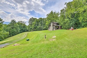 A log cabin with a stone foundation, surrounded by a lush green landscape and dense trees. - 'Turkey Hollow Cabin' w/ Stunning Open Views! (Clyde)