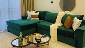 A living room with a green sectional sofa, two round mirrors, a small coffee table, and a large window with sheer curtains. - Tastefully furnished 2 bedroom apartment in  Kilimani (Nairobi)
