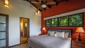 Une chambre à coucher avec un grand lit, deux lampes de chevet, une fenêtre donnant sur les arbres et un ventilateur de plafond.