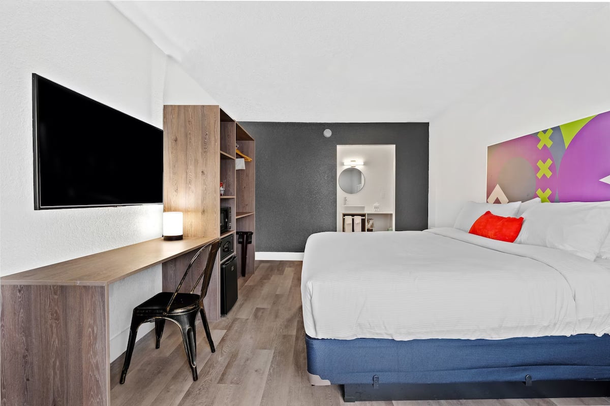 A modern hotel room with a bed, a desk, a chair, a TV, and a colorful wall decoration.
