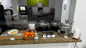 Montaje tipo bufé con diversos alimentos y bebidas, incluyendo un bol de naranjas, frascos con mermeladas y una máquina de café.