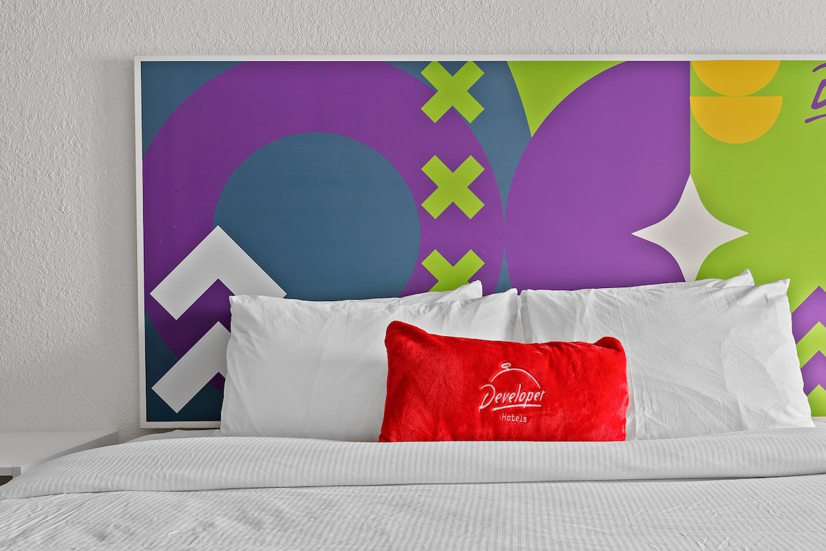 A hotel room with a bed, white bedding, a red pillow, and a colorful abstract wall art.