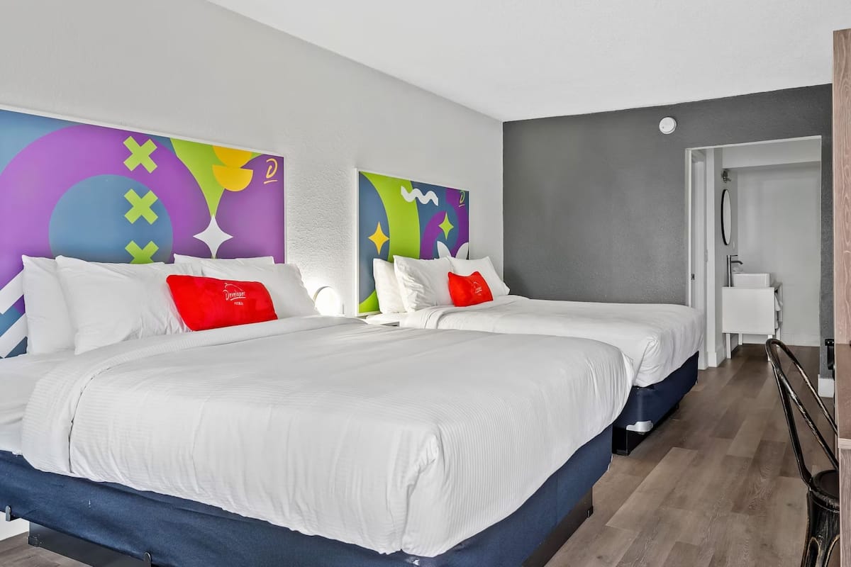 A hotel room with two beds, white bedding, colorful abstract wall art, a grey accent wall, and a wooden floor.
