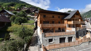 A wooden chalet with a stone base, multiple balconies, and a view of surrounding houses and mountains. - L'Étendard  appartement 7 (Vaujany)