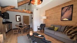 A cozy living room with a grey sofa, wooden walls, a dining area with a wooden table and chairs, and a kitchen with modern appliances. - L'Étendard  appartement 7 (Vaujany)