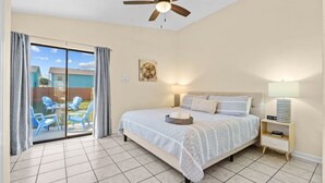 Une chambre avec un lit, un ventilateur de plafond, une table de chevet, une lampe et une vue sur un espace extérieur aménagé avec des chaises, accessible par une porte-fenêtre coulissante.