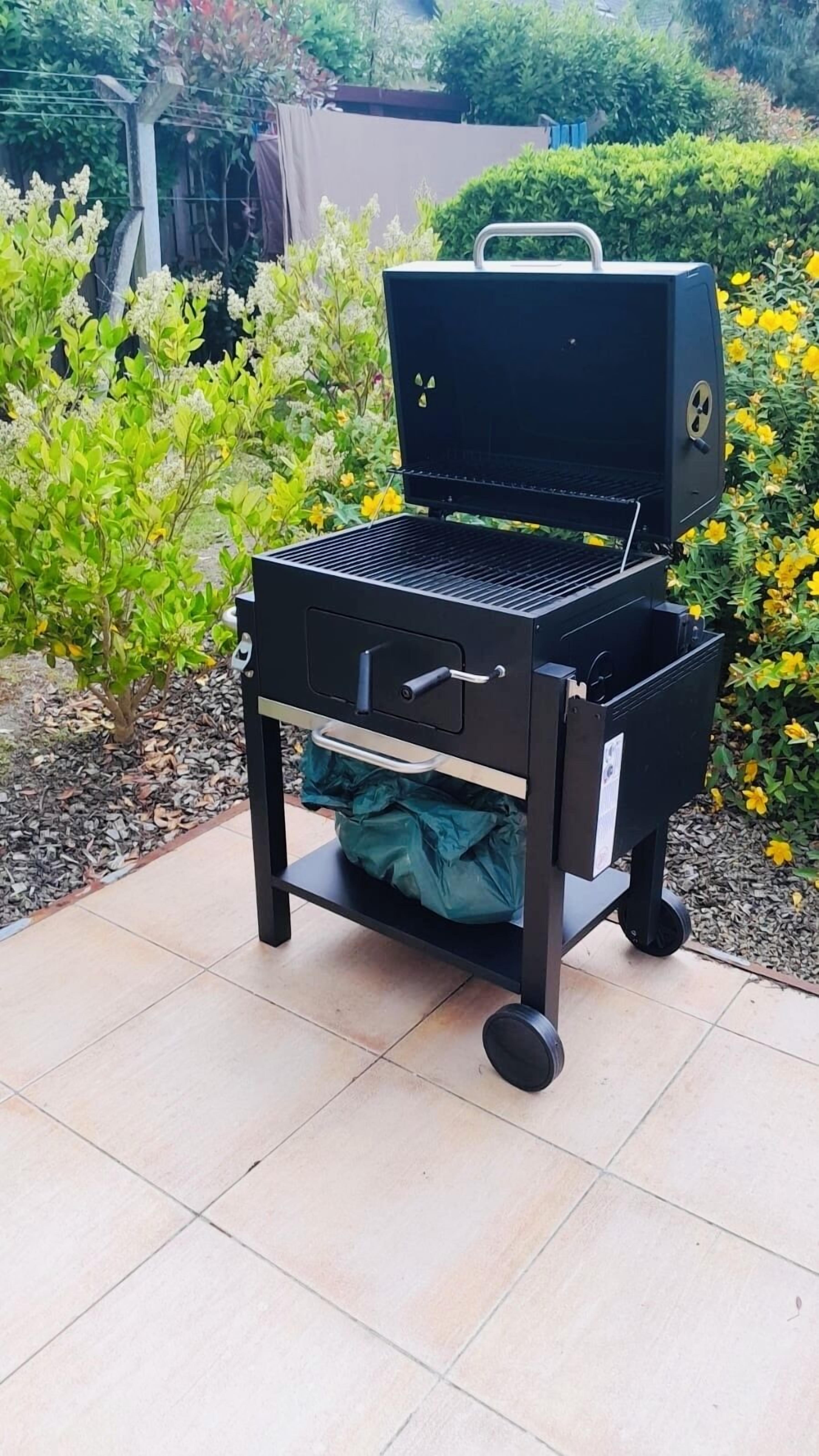 A portable barbecue grill with a storage drawer and wheels, placed on a tiled patio.