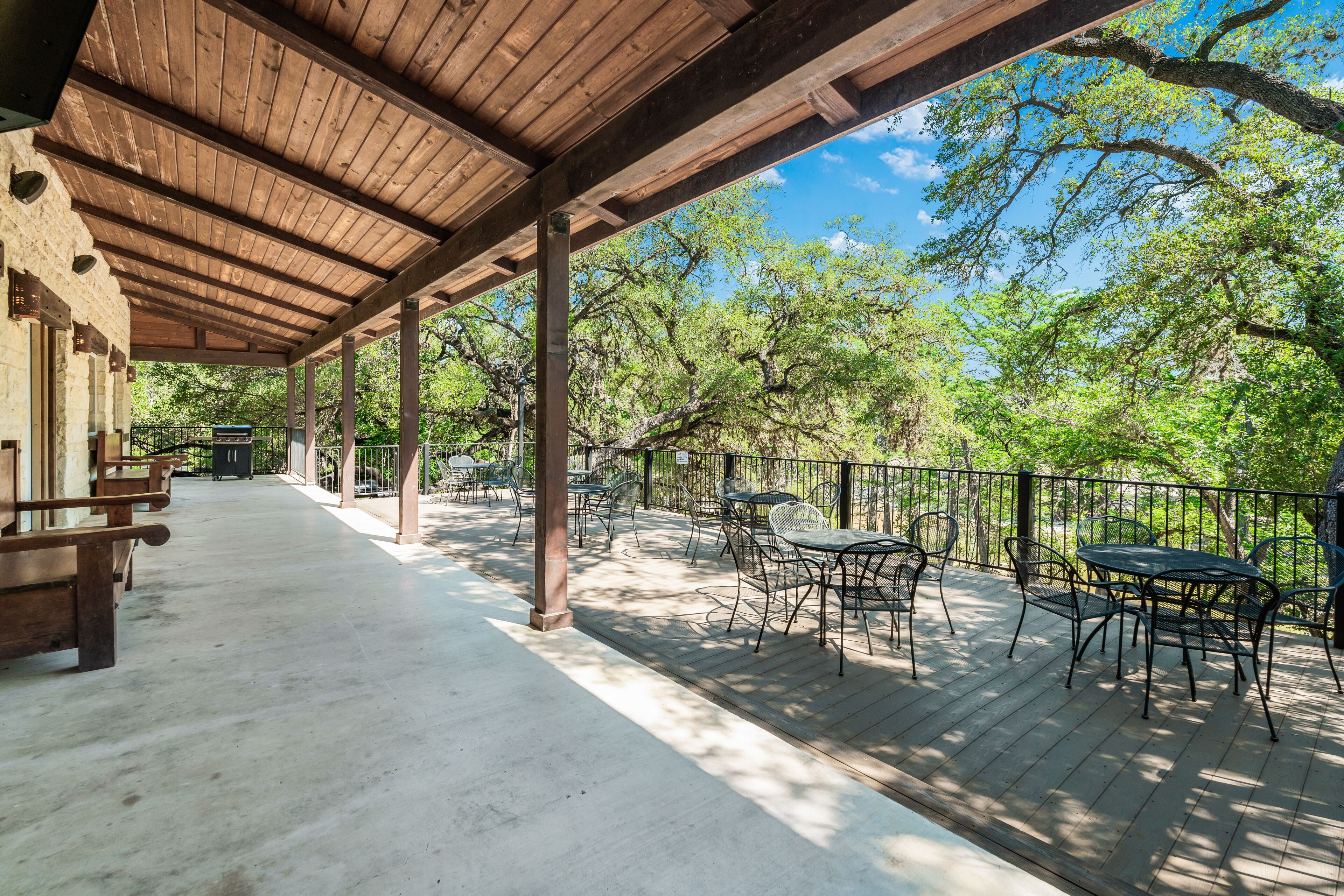 A covered outdoor patio with wooden benches and tables, surrounded by trees and a clear sky.