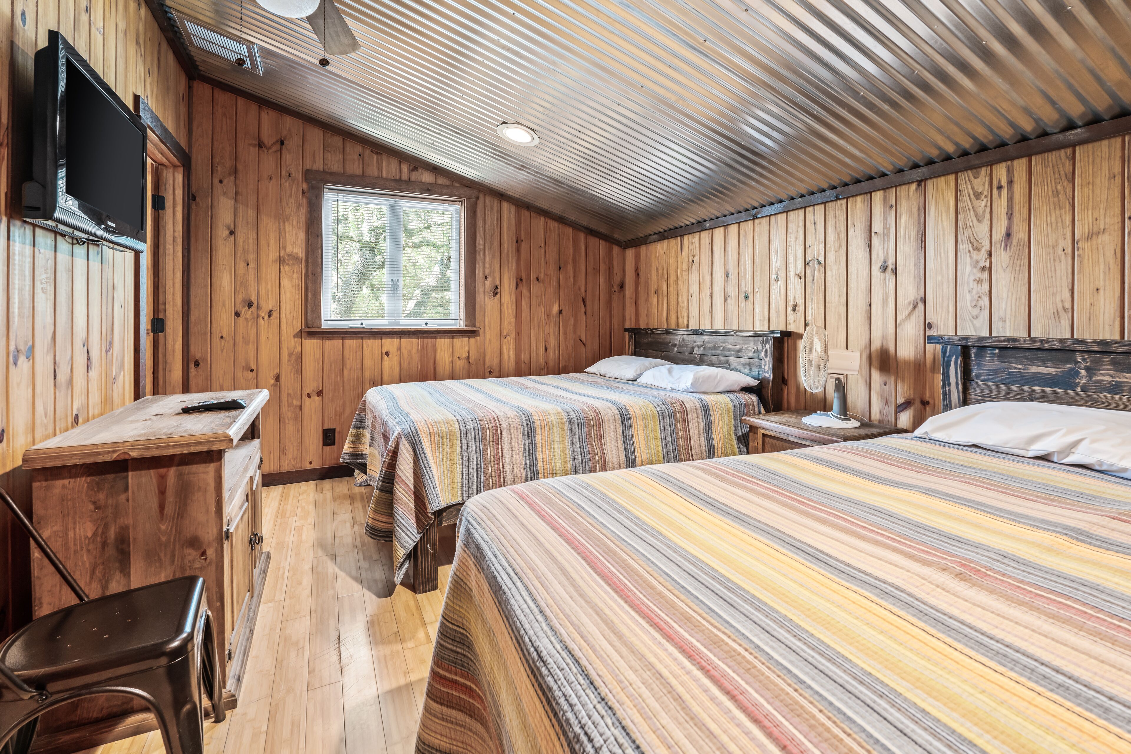 A wooden cabin room with two beds, a desk, a chair, and a television.