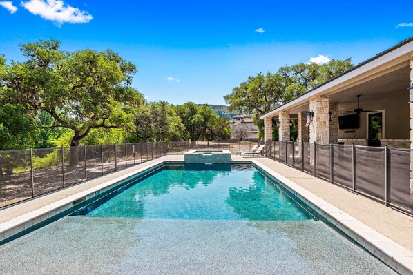 A rectangular swimming pool with a stone border, surrounded by a fence and trees.