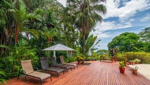 A wooden deck with lounge chairs and a table, surrounded by lush tropical plants and palm trees. - 4 bdrs, Casa Shanti, ocean view & large pool! (Savegre de Aguirre)