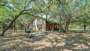 A rustic cabin with a stone exterior and a green roof, surrounded by trees and a leaf-covered ground. - Steps from THE Frio River with river access included, the perfect Concan cottage with the best price (Concan)