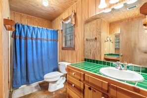 A wooden bathroom with a white toilet, a blue shower curtain, and a green-tiled sink. - Steps from THE Frio River with river access included, the perfect Concan cottage with the best price (Concan)