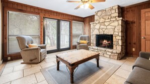 A cozy living room with a stone fireplace, two armchairs, a wooden coffee table, and large windows with blinds. - Directly ON THE Frio river, with a private pool and room for up to 37! (Concan)