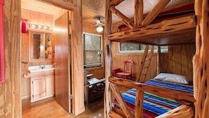 A rustic wooden cabin interior with a bunk bed, a small desk, a chair, a vanity with a mirror, and a window with blinds. - Cottage in the heart of Concan surrounded by nature & less than 5-minute drive to THE Frio River (Concan)
