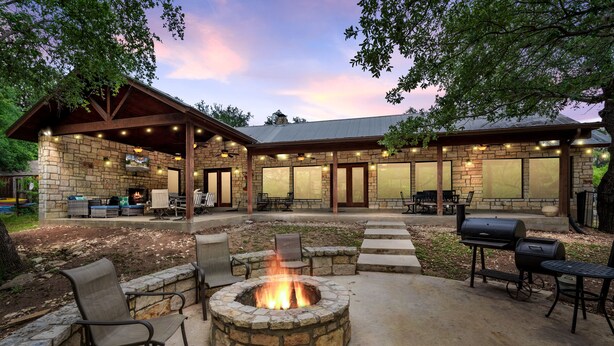 Un patio de piedra con un hogar, asientos exteriores y una zona cubierta con luces colgantes.