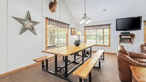 Een eetgedeelte met een houten tafel en banken, een opgehangen hertenkop, een sterdecoratie en een televisie aan de muur.