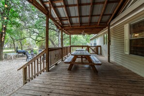 Une terrasse en bois avec des tables de pique-nique et une rambarde, donnant sur une cour arrière équipée d’un barbecue et de chaises.