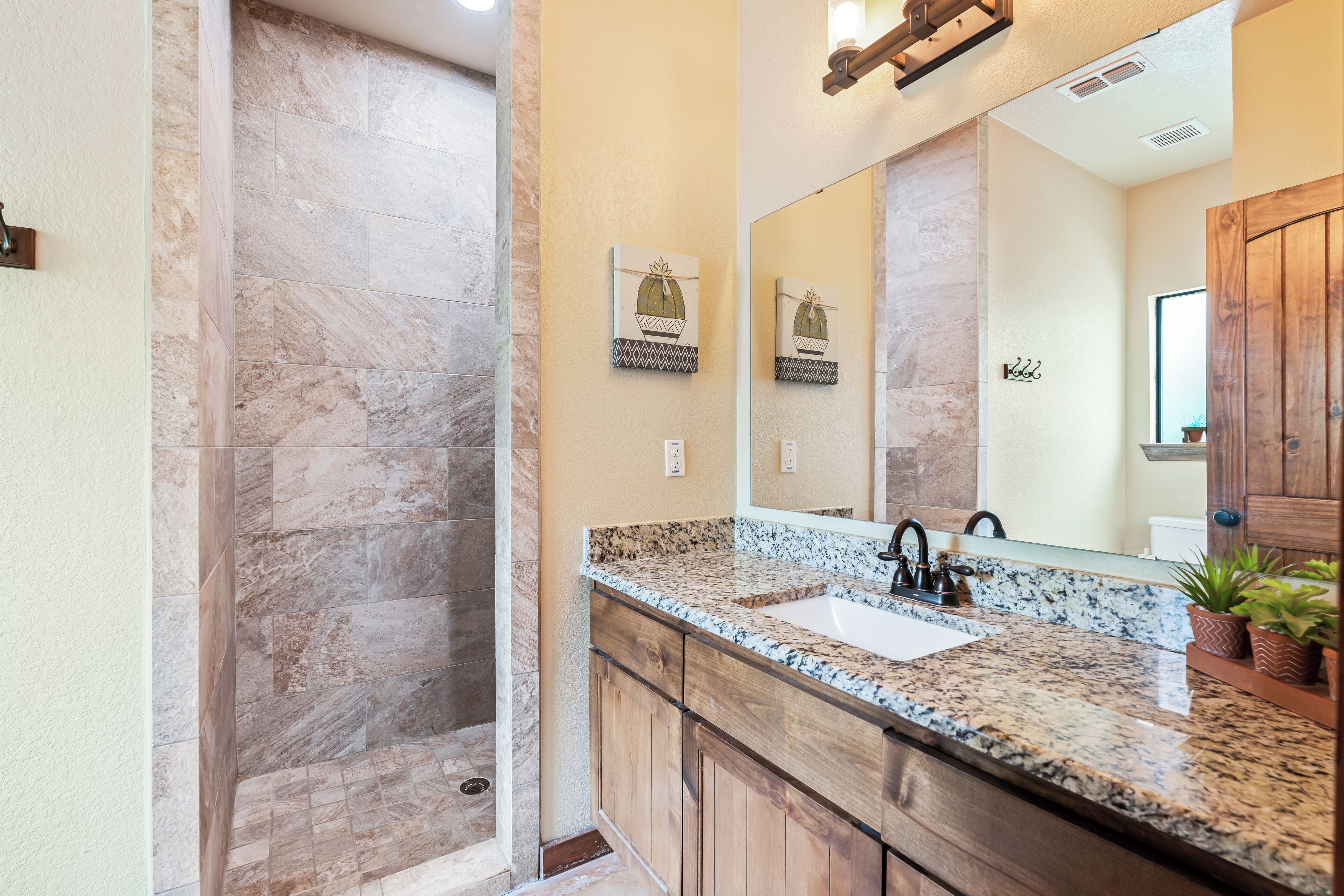 A bathroom with a marble shower, a granite countertop sink, wooden cabinets, and a large mirror.