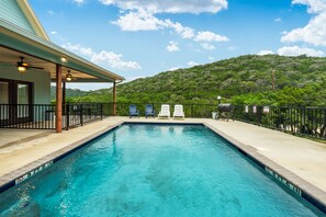 A swimming pool with a view of a green hill, lounge chairs, and a covered patio area.