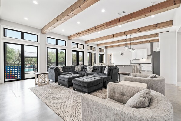 A spacious living room with a sectional sofa, ottomans, and a large rug. There are multiple windows providing natural light and a view of the outdoors.