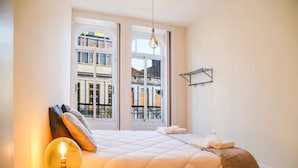 Une chambre à coucher avec un lit, une table de chevet avec une lampe et une fenêtre donnant sur des immeubles.