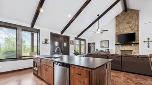 A spacious kitchen with a central island, wooden cabinets, and a dishwasher. There is a stone fireplace and a flat-screen TV mounted above it. - Private pool, volleyball, basketball, outdoor fireplace & less than 5-minute drive to THE Frio River (Concan)