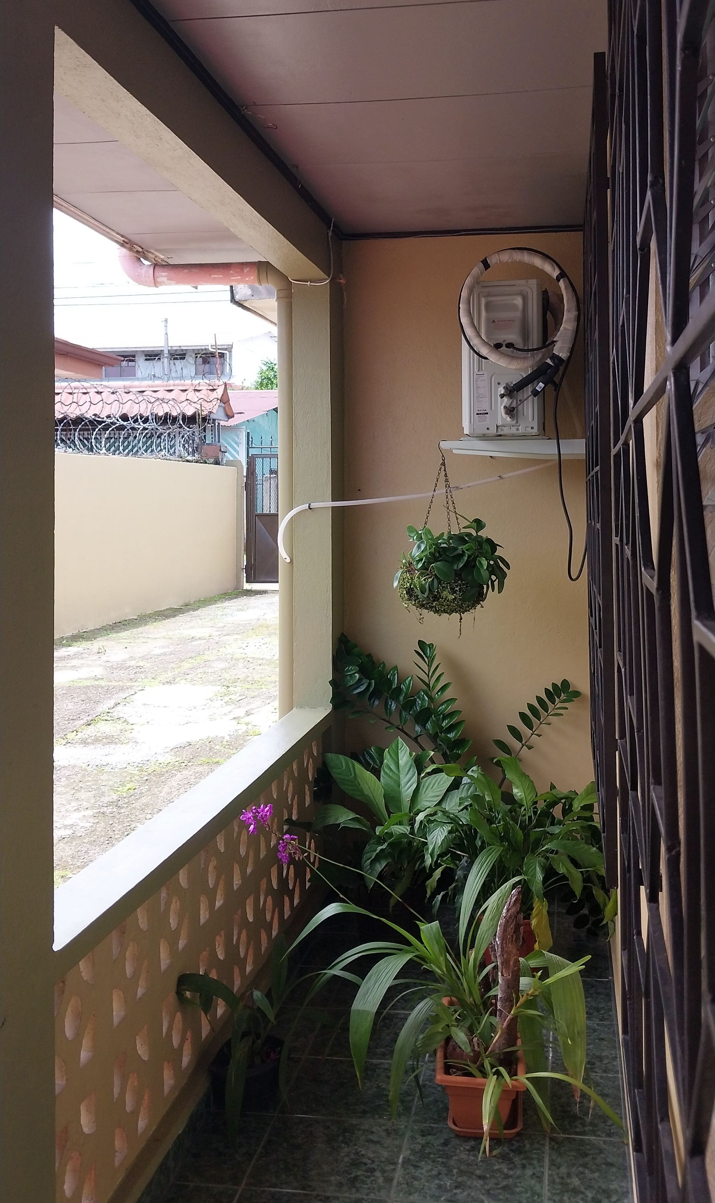 Un balcón pequeño y cerrado con suelo de baldosas, una ventana que ofrece vistas a un edificio y varias plantas en macetas.