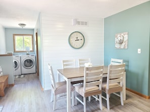Un comedor con una mesa y sillas de madera, un reloj en la pared y vistas a un cuarto de lavandería con lavadoras.
