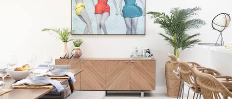 Uma sala de jantar moderna com uma mesa de madeira posta para dois, um banco e cadeiras de vime. Há uma pintura grande na parede acima da mesa, um vaso com planta e um globo em um suporte.