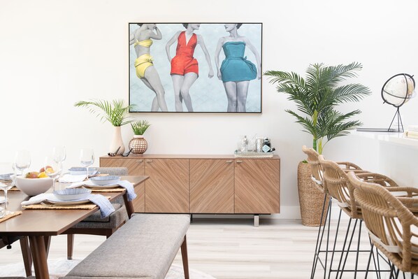 A modern dining area with a wooden dining table set for two, a bench, and wicker chairs. There is a large painting on the wall above the table, a potted plant, and a globe on a stand. - Luxury Home - Jacuzzi! Mini-Golf! Ping-Pong! Pets! 2 Desks! Yoga! Beach Gear! (North Palm Beach)