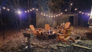 Un espace extérieur aménagé avec des chaises en bois et un foyer, entouré d’une clôture en bois et illuminé de guirlandes lumineuses.