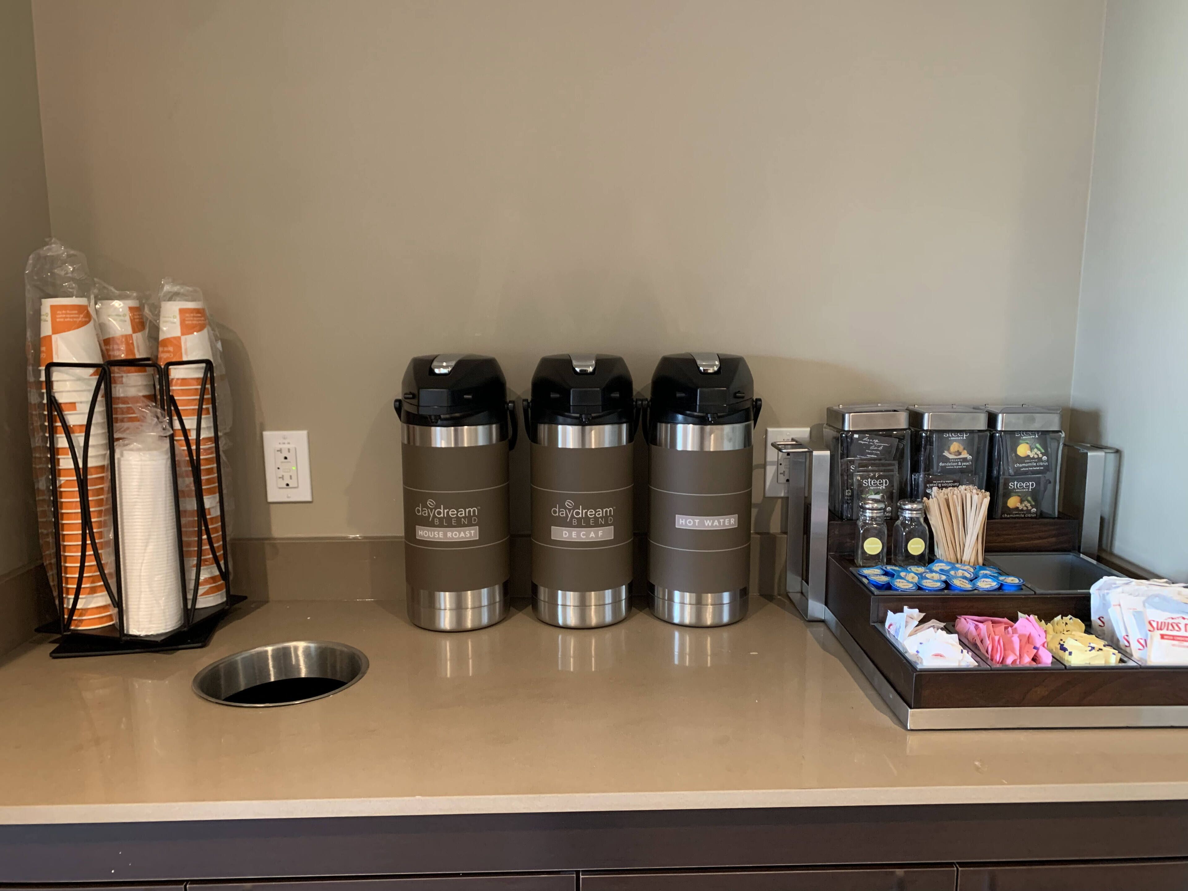 A coffee station with three thermos cups labeled 'daydream blend', 'house roast', and 'decaf'. There are also disposable cups, a coffee machine, and a tray with coffee pods and napkins.