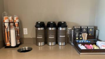 A coffee station with three thermos cups labeled 'daydream blend', 'house roast', and 'decaf'. There are also disposable cups, a coffee machine, and a tray with coffee pods and napkins.