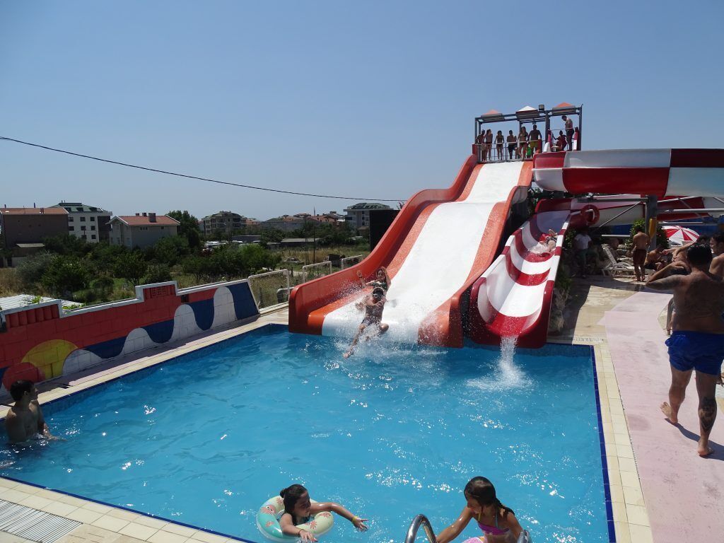 Una piscina con tobogán acuático, rodeada de una solarium y edificios al fondo.