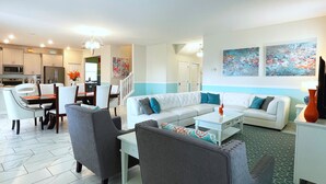 A modern living room with a sofa, chairs, a coffee table, and a dining area with chairs and a table. There is a kitchen in the background with appliances and cabinets.