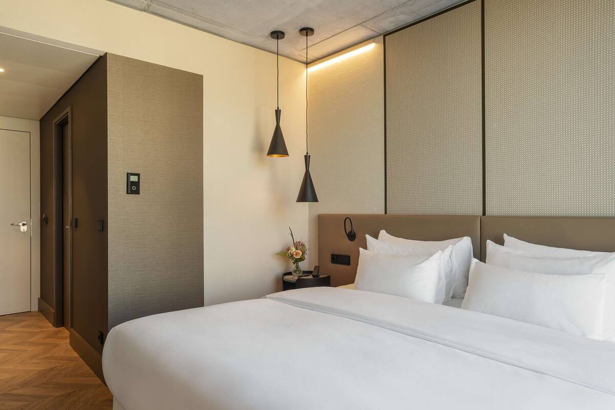 Een hotelkamer met een groot bed, een nachtkastje met lamp en een bedieningspaneel aan de muur.