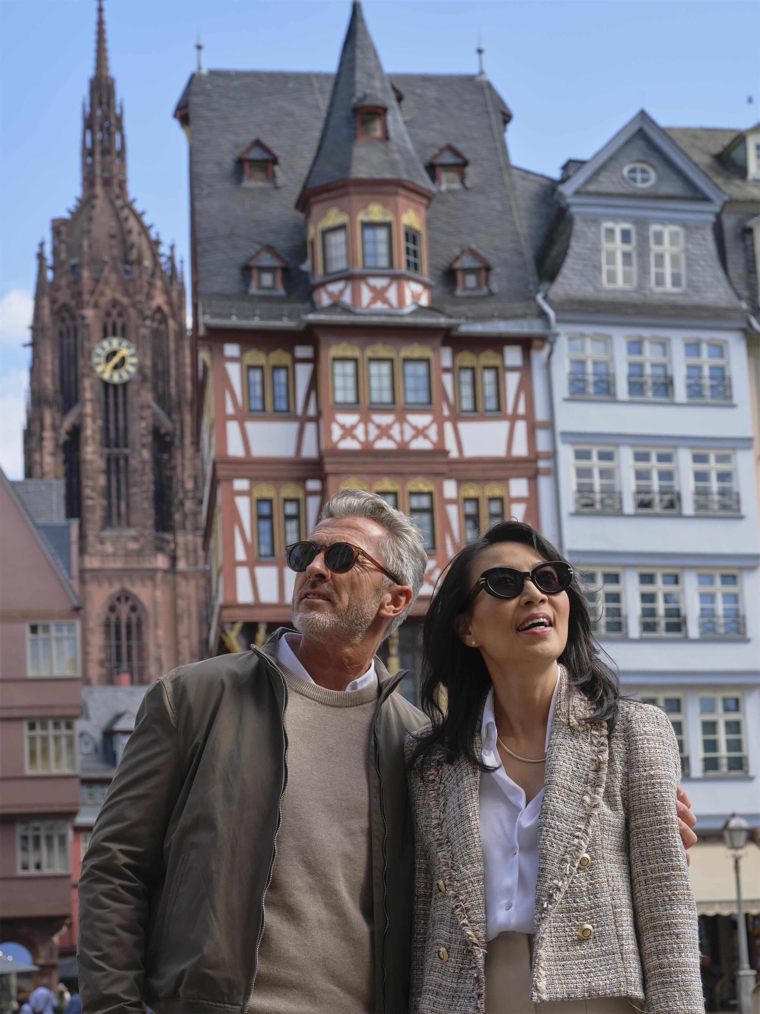 A couple standing in front of a historic building with a clock tower and traditional architecture.