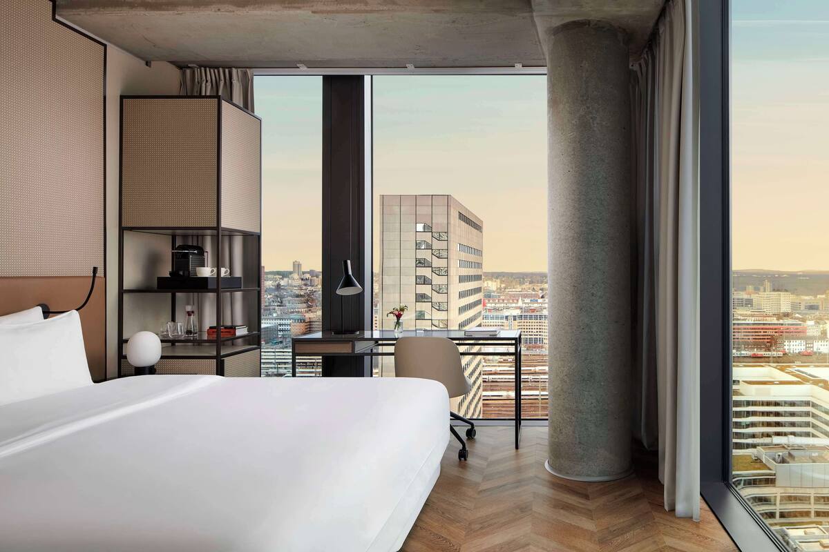 Een moderne hotelkamer met een groot bed, een bureau met stoel, en een uitzicht op de stad door grote ramen.