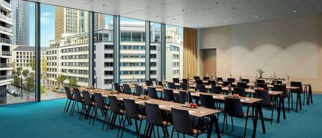 Un salón de conferencias con mesas y sillas, ventanas amplias con vista a la ciudad y piso alfombrado.