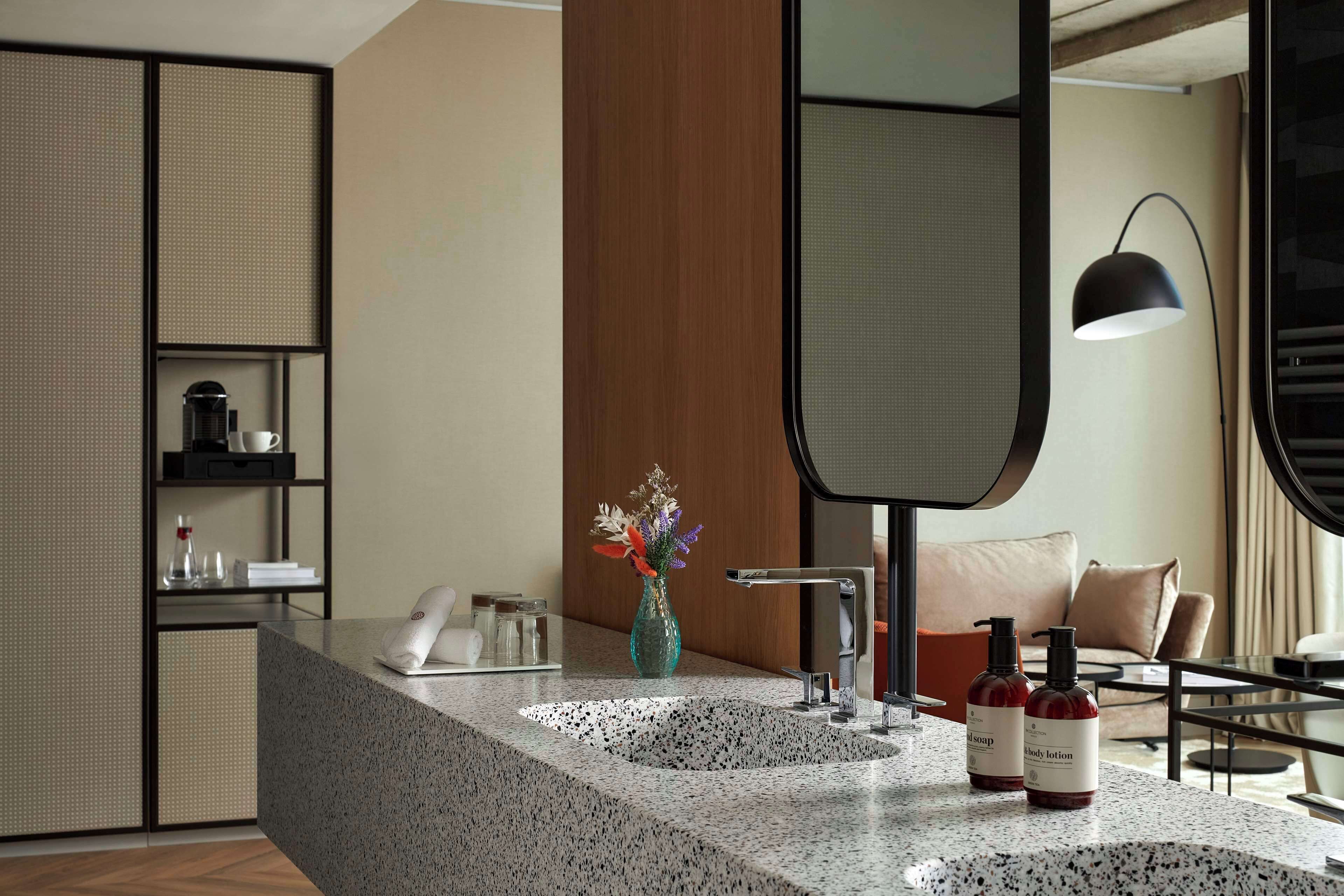 A modern bathroom with a granite countertop, two sinks, a mirror, a vase with flowers, and a coffee maker.