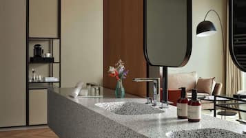 A modern bathroom with a granite countertop, two sinks, a mirror, a vase with flowers, and a coffee maker.