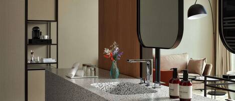 A modern bathroom with a granite countertop, two sinks, a mirror, a vase with flowers, and a coffee maker.