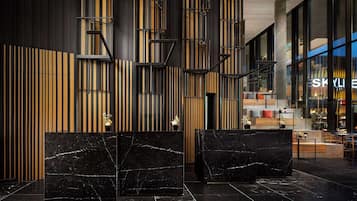 A modern lobby with black marble reception desks, vertical wooden slats, and a visible restaurant area in the background.