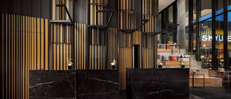A modern lobby with black marble reception desks, vertical wooden slats, and a visible restaurant area in the background.
