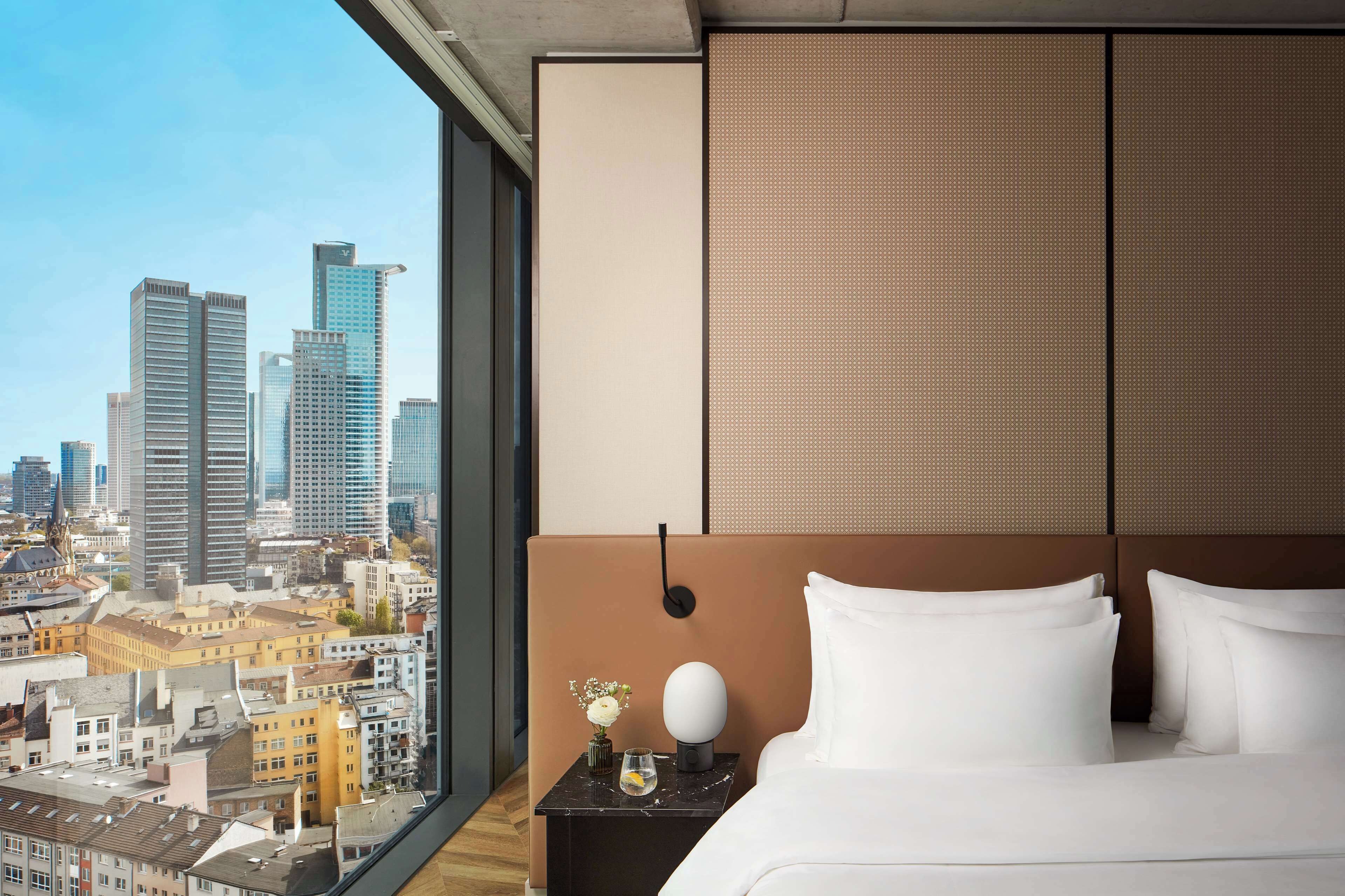 Ein Hotelzimmer mit großem Fenster und Blick auf eine Stadtlandschaft, ein Bett mit weißen Leinen, ein Nachttisch mit Blumen in einer Vase und ein modernes Kopfstück.